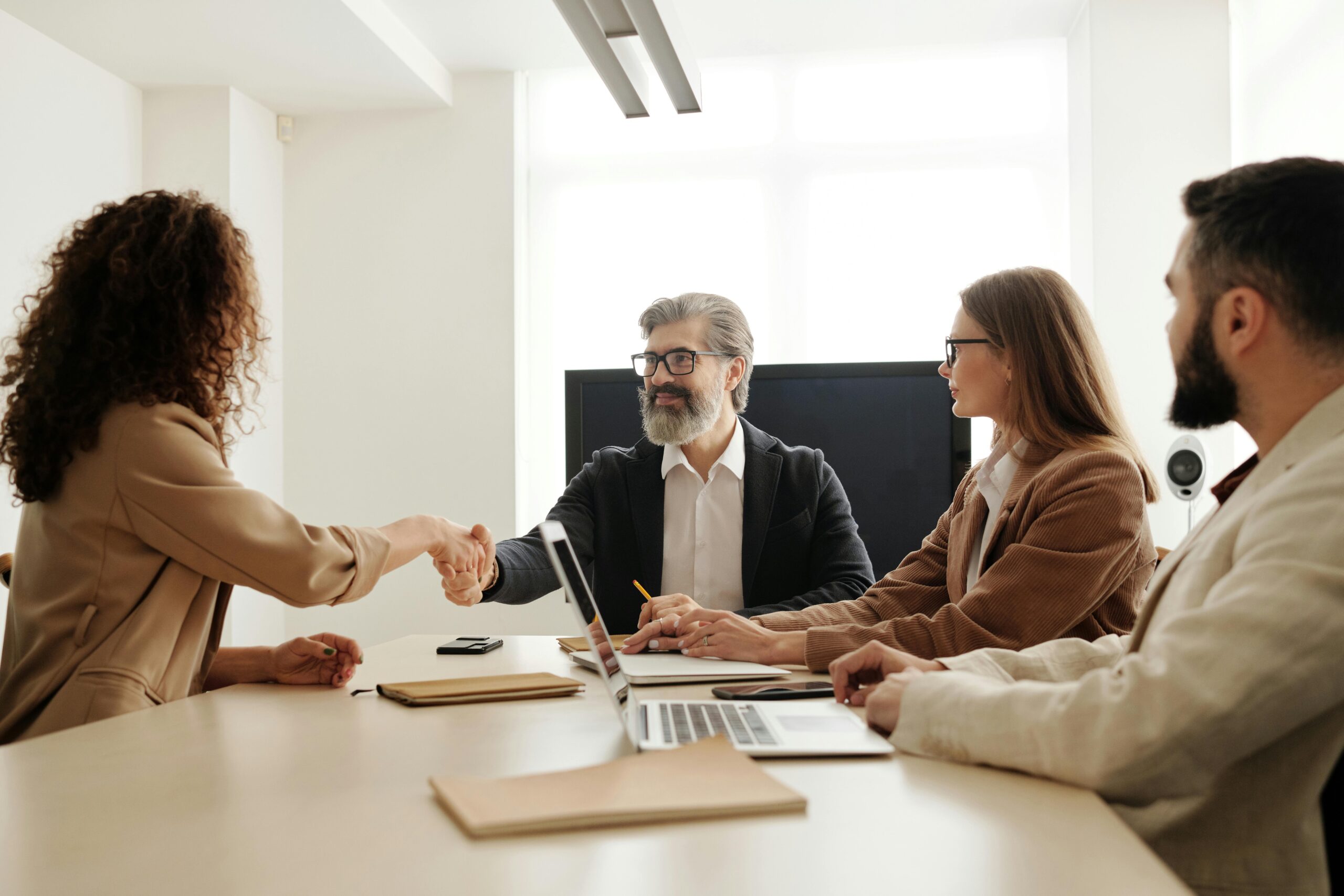 Executives shaking hands across conference table, sealing agreement as colleagues observe.