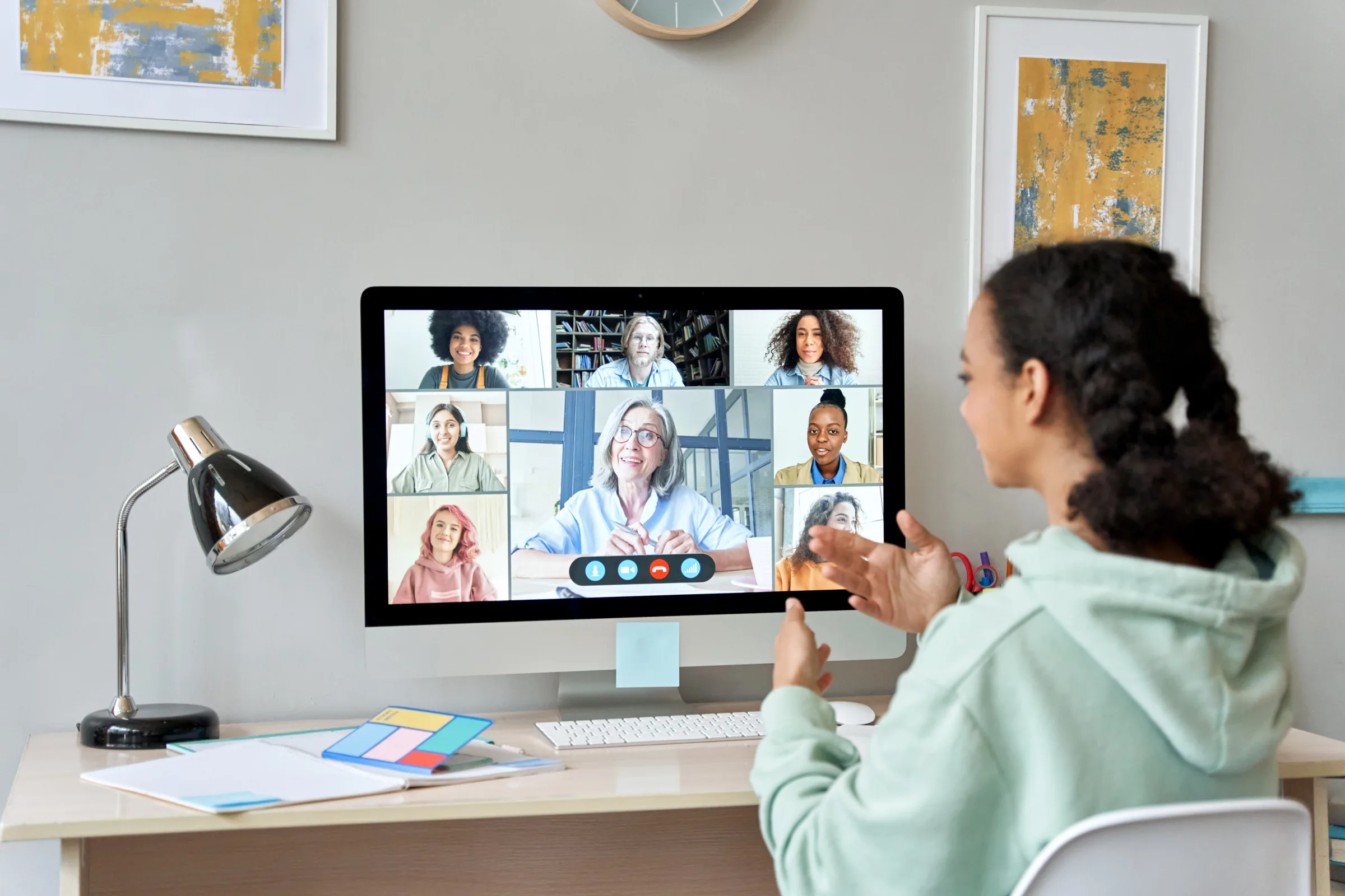 Young woman gesturing during remote meeting displayed on large desktop monitor.